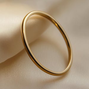 May include: A gold ring with a minimalist design. The circular ring is made of polished metal and sits on a soft, cream-coloured fabric. The contrast of textures and colours is striking. The ring is a piece of jewellery.