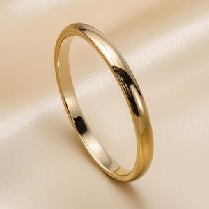 Könnte beinhalten: Ein goldener Bandring mit poliertem Finish. Der Ring ist vor einem cremefarbenen, satinartigen Hintergrund platziert. Die glatte Oberfläche des Rings reflektiert das Licht und erzeugt Glanzlichter und Schatten.