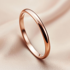 Puede incluir: Un anillo de oro rosa con una superficie lisa y pulida. El anillo se encuentra sobre una tela suave de color melocotón, creando una estética minimalista y elegante. El diseño del anillo es simple y clásico.