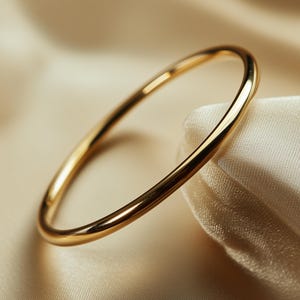 Puede incluir: Un brazalete de oro con una superficie lisa y pulida. El brazalete es circular y descansa sobre una tela color crema, creando un contraste de texturas. La joya es simple y elegante.