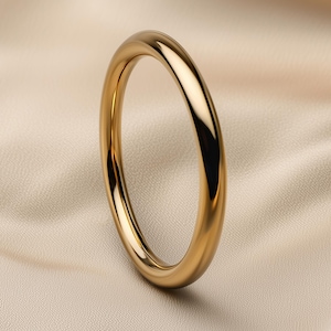 May include: A gold-coloured, rounded bangle bracelet. The smooth, polished surface reflects light, creating highlights and shadows. The bracelet is set against a neutral, textured background.