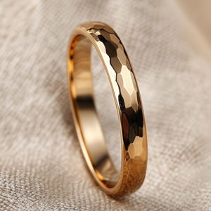 Puede incluir: Un anillo de oro con textura martillada. El anillo tiene una banda con pequeñas facetas irregulares que captan la luz. El anillo está sobre un fondo neutro, resaltando su brillo metálico. Es una pieza de joyería.