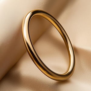 May include: A gold-coloured, oval-shaped bangle bracelet. The smooth, polished surface reflects light, creating highlights and shadows. The bracelet is set against a backdrop of a soft, beige fabric.