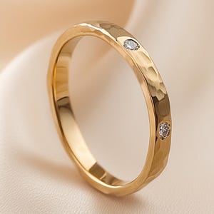 Può includere: Un anello d'oro con una texture martellata e due piccole gemme trasparenti. L'anello è posto su uno sfondo morbido e neutro, che ne evidenzia la superficie lucida e riflettente. Il design dell'anello è semplice ed elegante.