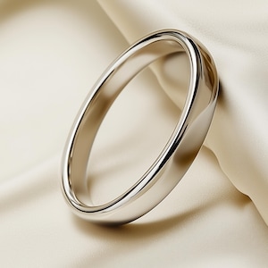 May include: A polished silver bangle bracelet. The smooth, rounded band reflects light, creating highlights and shadows. The bracelet is set against a soft, off-white fabric background, enhancing its metallic sheen. A simple, elegant piece of jewellery.