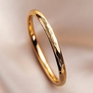 Puede incluir: Un brazalete de metal dorado martillado. El brazalete tiene una superficie texturizada con una serie de pequeñas hendiduras. El brazalete está sobre un fondo suave y neutro.