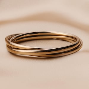 May include: A gold-colored bangle bracelet with three interlocking bands. The smooth, polished metal reflects light, creating highlights and shadows. The bracelet is set against a neutral, cream-colored background.