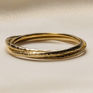 May include: A set of three gold-colored rings interlocked, creating a textured, hammered effect. The rings are displayed on a light-colored, slightly textured surface. The jewelry has a simple, elegant design.