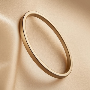 Könnte beinhalten: Ein goldfarbenes, oval geformtes Armreif-Armband. Das Armband ist vor einem beigefarbenen, strukturierten Hintergrund platziert. Der Armreif ist glatt und hat ein schlichtes, elegantes Design, das für den Alltag geeignet ist.