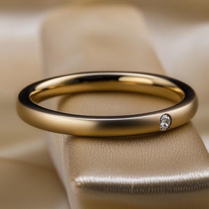 May include: A gold ring with a single diamond. The ring is displayed on a neutral-toned surface, with a soft, blurred background. The ring's band is smooth and polished, and the diamond is set flush with the band.
