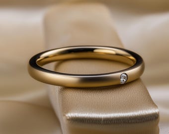 Könnte beinhalten: Ein goldfarbener Ring mit einem einzelnen, kleinen Diamanten. Der Ring liegt auf einer neutral getönten Oberfläche mit einem weichen, verschwommenen Hintergrund. Der Ring ist glatt und poliert, der Diamant bündig eingesetzt.