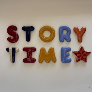 May include: A set of ten individual ceramic letters and a star shape arranged to spell STORY TIME. The letters are glossy and colourful, featuring red, dark blue, yellow, light blue, orange, and a striped pattern on the letter T and star.
