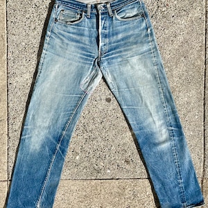 Puede incluir: Un par de pantalones vaqueros azules con un desgarro reparado en la pierna izquierda. Los vaqueros tienen un aspecto descolorido y están colocados sobre una superficie de hormigón.