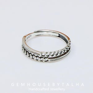 Puede incluir: Un anillo ajustable de plata con diseño de cadena. El anillo está marcado con "5925" y presenta el texto "GEMHOUSEBYTALHA Handcrafted Jewellery" en la parte inferior. El anillo tiene un acabado pulido y está sobre un fondo blanco.