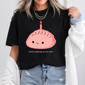 Może przedstawiać: Czarny t-shirt z grafiką różowego pierożka z uśmiechniętą buzią i zapaloną świeczką urodzinową na górze. Poniżej pierożka znajduje się napis "CUTEST DUMPLING AT THE PARTY" wykonany białymi wielkimi literami, wyśrodkowany na wysokości klatki piersiowej.