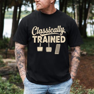 Könnte beinhalten: Schwarzes T-Shirt mit dem Text "Classically Trained" in abgenutzter Schrift, zusammen mit Abbildungen von Autopedalen. Das Shirt hat einen Rundhalsausschnitt und scheint aus einem weichen Material zu sein.