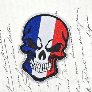 Peut inclure: Un écusson brodé représentant un crâne humain aux couleurs du drapeau français, avec des bandes verticales bleu, blanc et rouge, bordé de fil noir avec des détails de couture visibles sur les dents et les orbites oculaires.