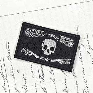 Peut inclure: Un écusson rectangulaire noir brodé représentant un crâne blanc au centre, flanqué de deux paires d'ailes blanches. Le texte MEMENTO est brodé au-dessus du crâne et MORI est brodé en dessous du crâne avec du fil blanc.