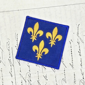 Può includere: Una toppa in tessuto di forma quadrata con uno sfondo blu intenso e tre simboli di giglio ricamati in giallo dorato disposti a triangolo. La toppa presenta un bordo cucito con un filo blu in tinta.