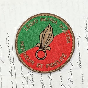 Op de afbeelding: Een ronde geborduurde patch met een gespleten groene en rode achtergrond en een goudkleurig granaatembleem in het midden. De tekst LEGIO PATRIA NOSTRA en HONNEUR ET FIDÉLITÉ is met gouddraad langs de rand van de patch geborduurd.