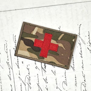 May include: A rectangular patch featuring a camouflage pattern in shades of green, brown, and tan. The patch has a dark olive green border and a prominent, embroidered red cross centered on the fabric, designed for attachment to gear or clothing.