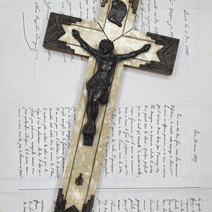 May include: An antique cross with a dark metal corpus on a cream-colored base, framed by dark metal and a starburst design. The cross is set against a backdrop of handwritten documents and greenery.