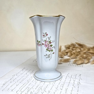 Pode incluir: Um vaso de porcelana branca com borda ondulada e canelada, com acabamento em dourado. O corpo apresenta uma delicada ilustração botânica de flores de macieira cor-de-rosa com folhas verdes e pequenos botões vermelhos sobre uma base dourada.