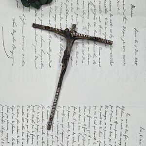 Peut inclure: Un crucifix antique détaillé avec une figure de Jésus-Christ, en métal sombre. La croix est posée sur une lettre manuscrite, avec une branche de feuilles vertes en arrière-plan.