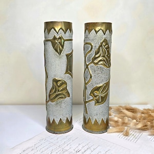 Peut inclure: Deux vases cylindriques en laiton avec une finition argentée texturée sur le corps. Chaque vase est orné de motifs floraux et de feuilles dorés en relief, avec des bords dentelés en forme de dents de scie en haut et en bas du métal.