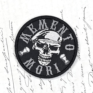 Op de afbeelding: Een ronde zwarte geborduurde patch met een witte schedel die een baret draagt in het midden. De tekst MEMENTO MORI is in witte hoofdletters rond de schedel geborduurd, met twee kleine witte bot-iconen die de woorden scheiden.