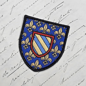 Peut inclure: Écusson brodé bleu et or avec un motif de bouclier. Le bouclier est orné de fleurs de lys dorées sur fond bleu. Un petit écusson rayé bleu et or est au centre, bordé de rouge et de noir.