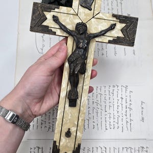 May include: An antique crucifix with a bronze Christ figure and decorative accents. The cross features a cream-colored, marbled design with bronze-colored geometric details. A small plaque with text is visible at the top.