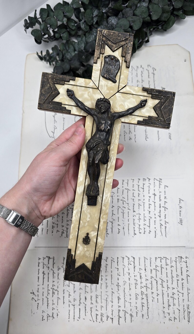 May include: An antique crucifix with a bronze Christ figure and decorative accents. The cross features a cream-colored, marbled design with bronze-colored geometric details. A small plaque with text is visible at the top.