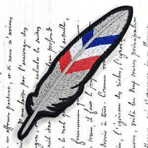 Peut inclure: Écusson brodé en forme de plume, argenté avec des rayures rouges, blanches et bleues. Le patch a une bordure noire et est probablement destiné à la décoration, peut-être pour des vêtements ou des accessoires.