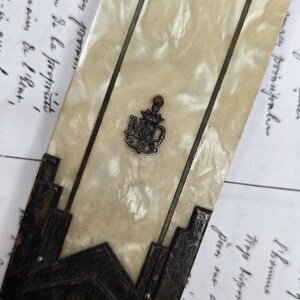 May include: An antique, rectangular mother-of-pearl and metal object with a decorative emblem. The object has dark metal accents and a central emblem with the letters "NMD". The background features handwritten script.