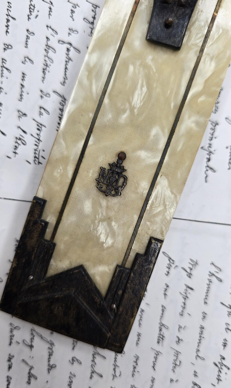 May include: An antique, rectangular mother-of-pearl and metal object with a decorative emblem. The object has dark metal accents and a central emblem with the letters "NMD". The background features handwritten script.