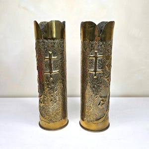 May include: A pair of tall, cylindrical brass vases featuring a textured, stippled surface finish. Each vase displays a prominent embossed cross symbol and decorative leaf patterns. The top edges are scalloped, and the base of each vessel is flat.