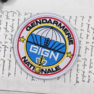 Op de afbeelding: Een ronde patch met de woorden "GENDARMERIE NATIONALE" en "GIGN" in een blauw en wit ontwerp. De patch heeft een witte rand met rode en gele accenten en twee gele sterren.