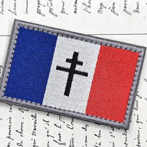 May include: Embroidered patch with the French flag design and a black Cross of Lorraine. The patch features blue, white, and red vertical stripes, with a grey border.