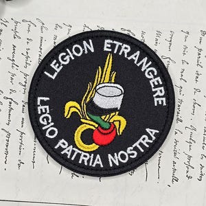 May include: A black, circular patch with white lettering that reads "LEGION ETRANGERE" and "LEGIO PATRIA NOSTRA". The patch features a central emblem with a cup, flames, and a red fruit. The patch is on a background of handwritten script.