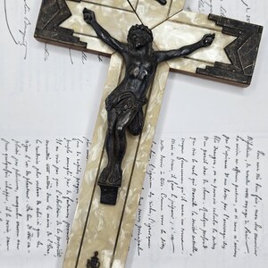 May include: Antique crucifix with a dark metal Christ figure. The cross features a cream-colored inlay with dark geometric accents. The background includes handwritten documents.