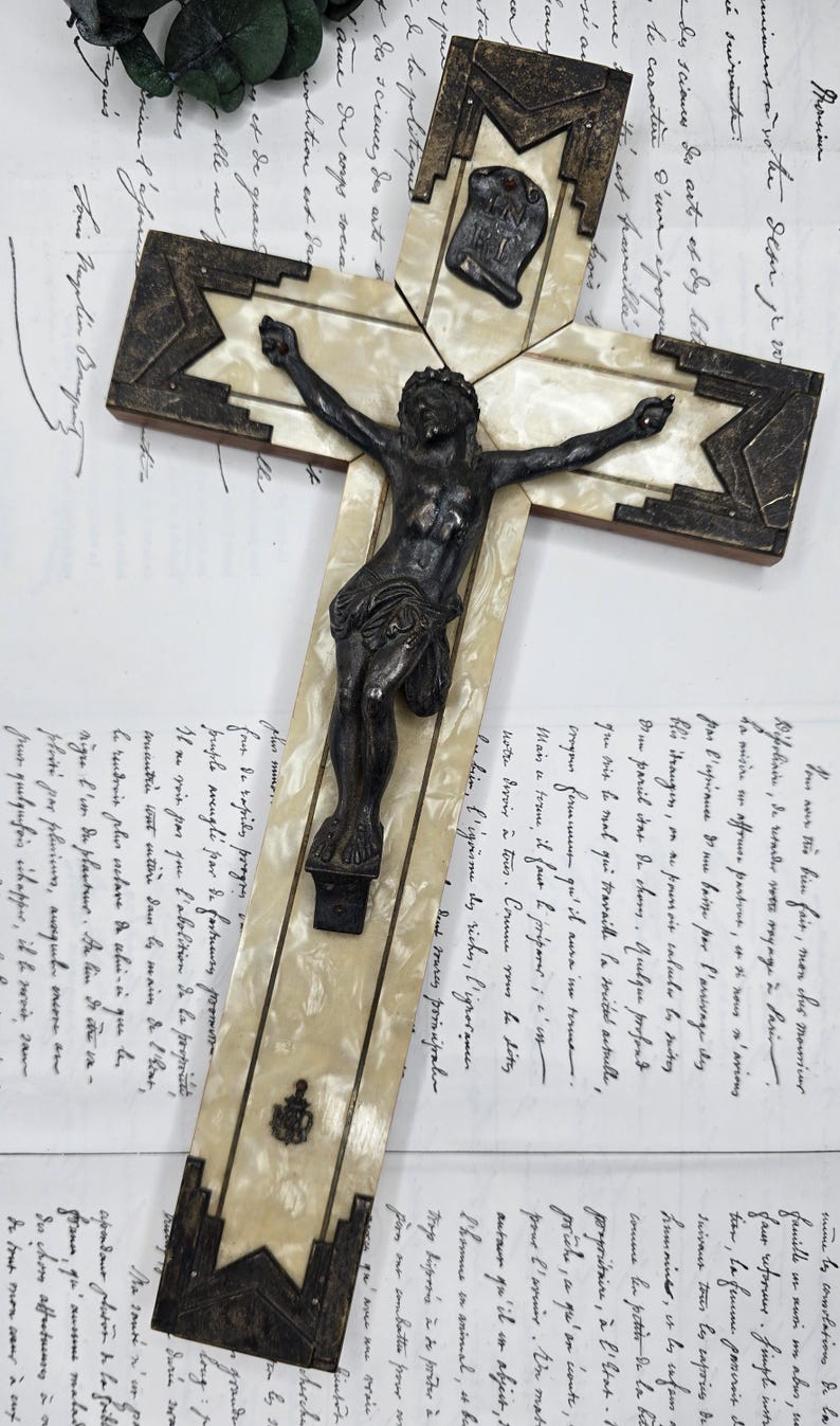 May include: Antique crucifix with a dark metal Christ figure. The cross features a cream-colored inlay with dark geometric accents. The background includes handwritten documents.
