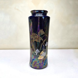 Può includere: Un vaso cilindrico in ceramica con una profonda smaltatura blu scuro iridescente. La superficie presenta un'illustrazione botanica dettagliata di fiori di iris dorati e bianchi, foglie verdi e un piccolo uccello, con una finitura riflettente.