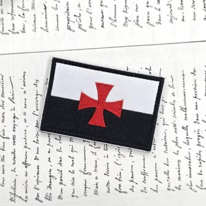 Puede incluir: Parche bordado con un diseño de bandera en negro, blanco y rojo. El parche tiene una sección superior blanca, una sección inferior negra y una cruz roja en el centro. El parche es rectangular y adecuado para ropa o accesorios.