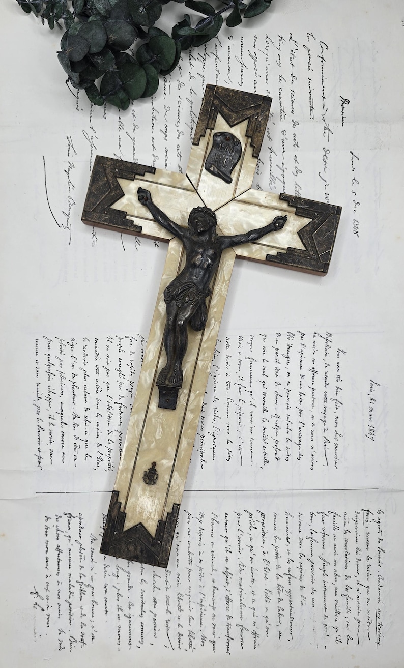 May include: An antique cross with a dark metal corpus on a cream-colored base, framed by dark metal and a starburst design. The cross is set against a backdrop of handwritten documents and greenery.