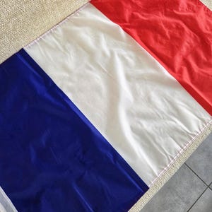Peut inclure: Un drapeau français avec des bandes verticales rouge, blanc et bleu. Le drapeau est fait d'un matériau brillant et léger. Les couleurs sont vives et bien distinctes. Le drapeau est posé sur une surface beige.