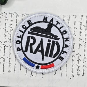 May include: A round white patch with the words "POLICE NATIONALE RAID" in black. The patch features a black emblem and a blue, white, and red stripe at the bottom. The patch is on a vintage paper background.
