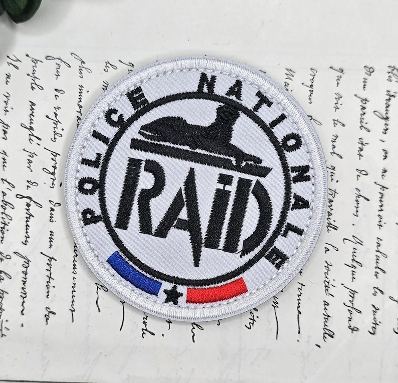 May include: A circular white patch with the words "POLICE NATIONALE RAID" in black. The patch has a black emblem and a blue, white, and red stripe at the bottom. The patch is set against a vintage paper background.