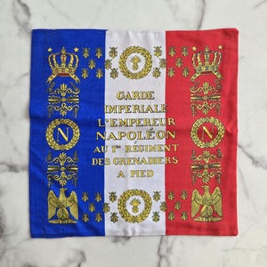 Könnte beinhalten: Ein rechteckiger Stoff mit einem vertikalen Streifenmuster in Blau, Weiß und Rot. Die blauen und roten Streifen weisen goldene Embleme, Kronen, Bienen und Text auf. Der weiße Streifen trägt den Text "GARDE IMPERIALE L'EMPEREUR NAPOLÉON AU 1er REGIMENT DES GRENADIERS A PIED."