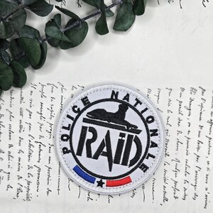 May include: A circular patch with the text "POLICE NATIONALE RAID". The patch is white with black text and a black emblem. It has a blue, white, and red stripe at the bottom. The patch is on a vintage paper background.